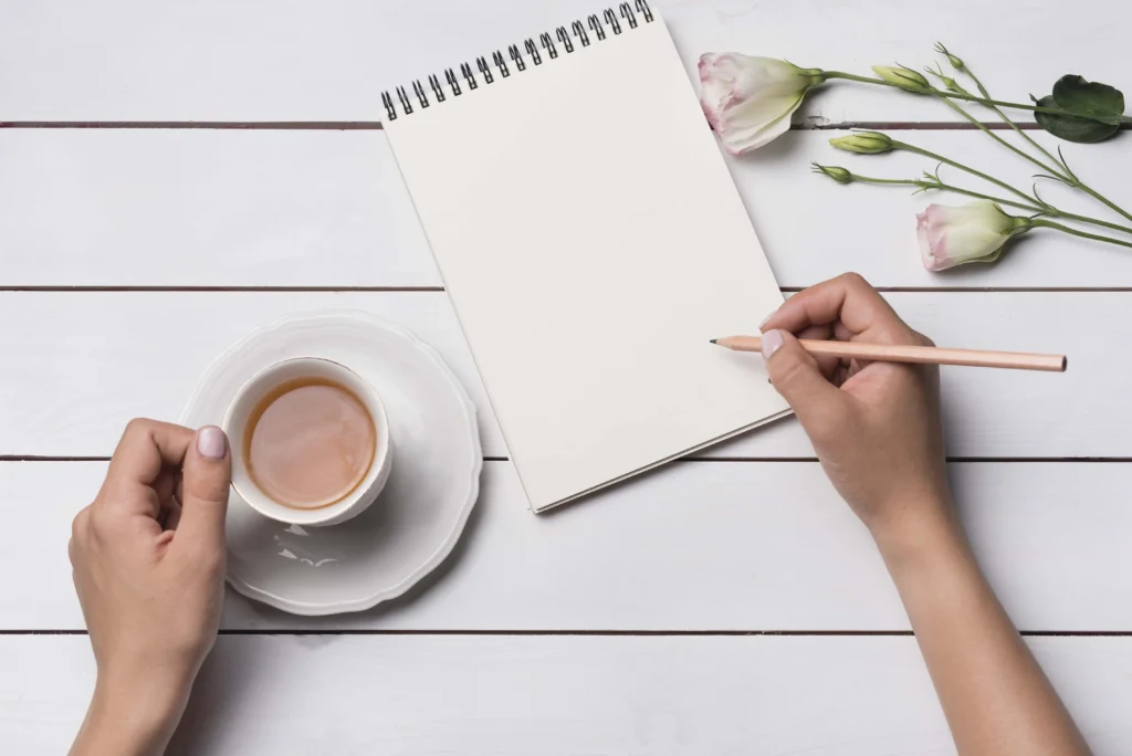 A close-up of a person writing in a notepad with a cup of tea on a wooden desk, capturing a daily gratitude practice.